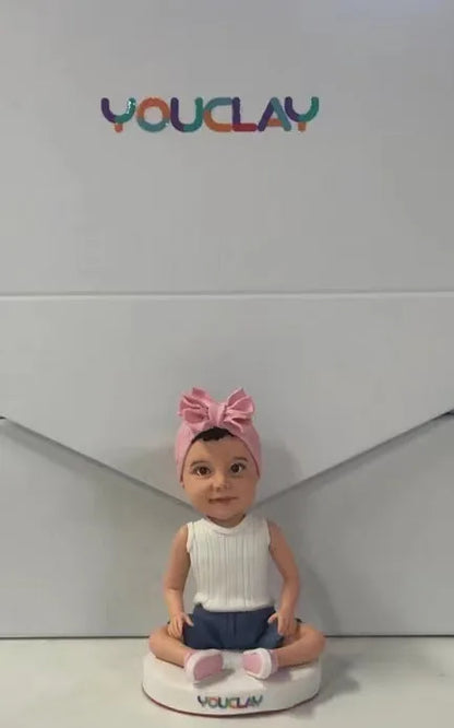 Seated baby girl in a big pink headband bow and white top, sculpted in polymer clay
