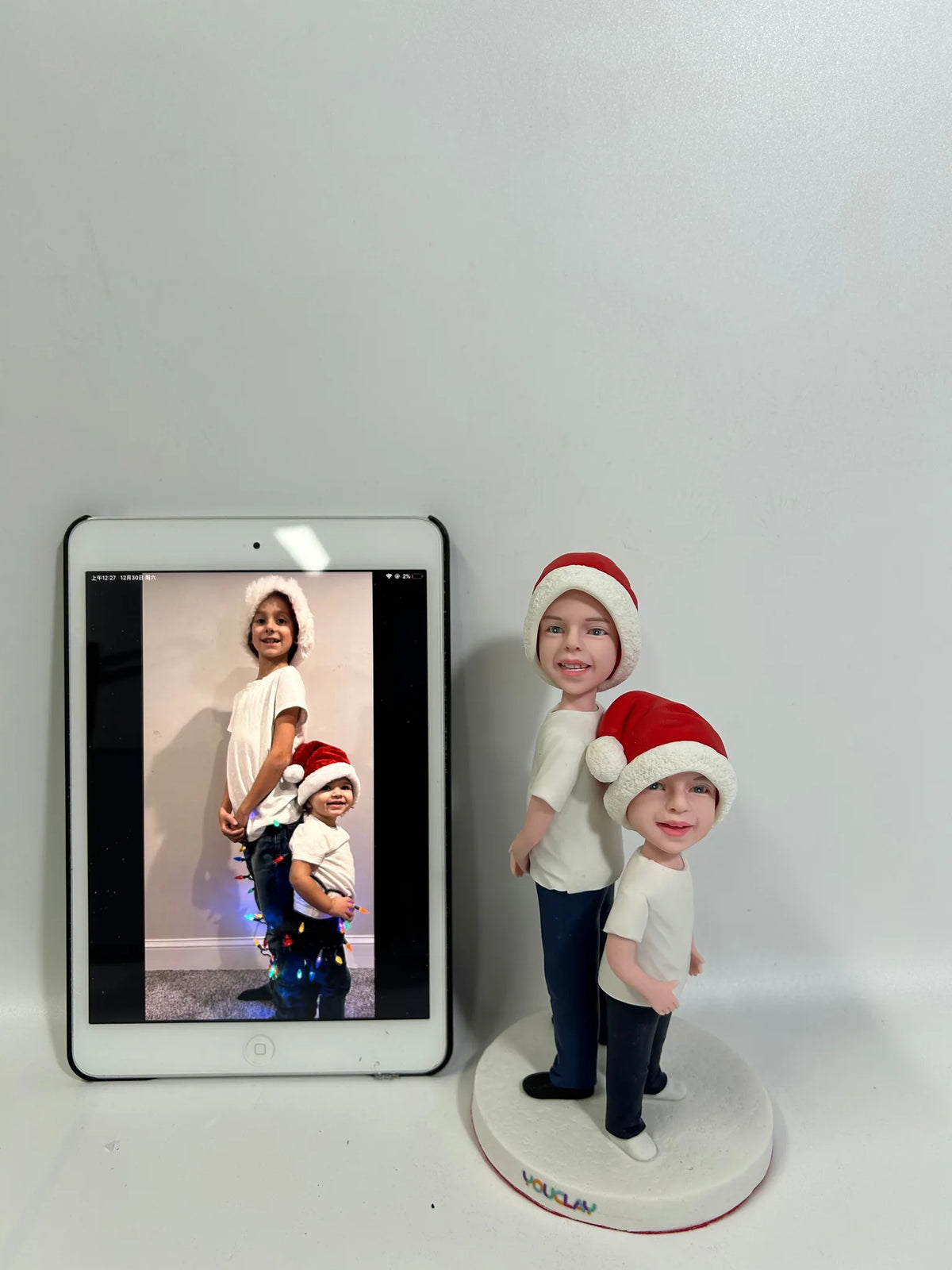 Two boys in Santa hats as clay figurines, standing beside a tablet showing their holiday photo